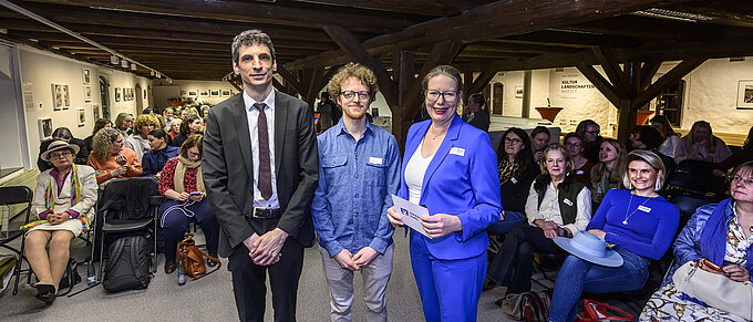 Drei Personen posieren vor einem Publikum in einem Raum mit Holzbalken. Im Hintergrund sitzt ein weibliches Publikum.