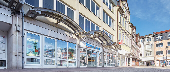 Straßenansicht mit Volksbank-Filiale und kik-Geschäft im Erdgeschoss eines mehrstöckigen Gebäudes bei blauem Himmel.