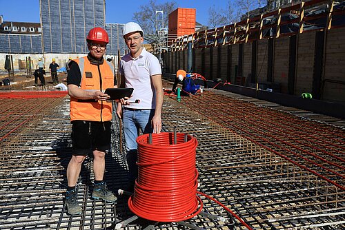 Bauarbeiter auf Baustelle mit Helm und Tablet vor Stahlkonstruktion, rote Kabelspule im Vordergrund.