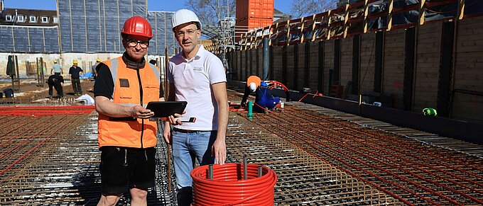 Bauarbeiter auf Baustelle mit Helm und Tablet vor Stahlkonstruktion, rote Kabelspule im Vordergrund.