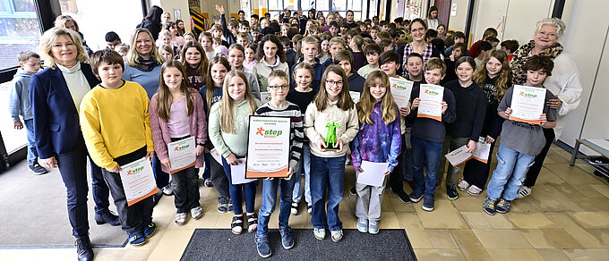 Große Gruppe von Kindern in einem Schulflur mit Zertifikaten und einem grünen Pokal, begleitet von Erwachsenen.