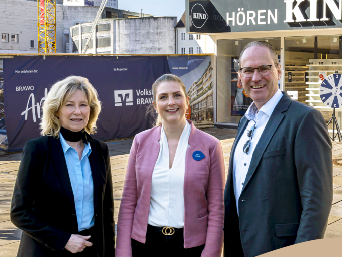 Drei Personen stehen vor den Brawo Arkaden in Wolfsburg, lächelnd in die Kamera, mit Bauarbeiten im Hintergrund.