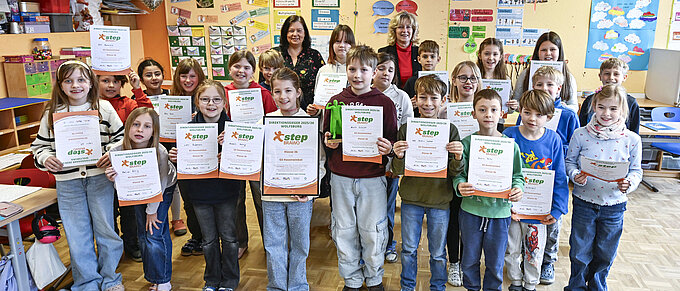 Kinder in einem Klassenzimmer halten stolz Zertifikate und posieren lächelnd, Erwachsene stehen dahinter.