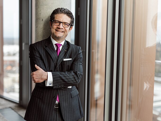 Mann im Anzug mit Krawatte steht lächelnd an einem Fenster, Arme verschränkt.