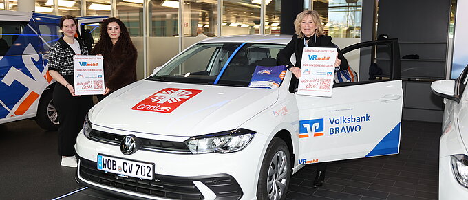 Drei Personen präsentieren ein VRmobil der Volksbank Brawo und Caritas mit Plakaten in der Autostadt in Wolfsburg.