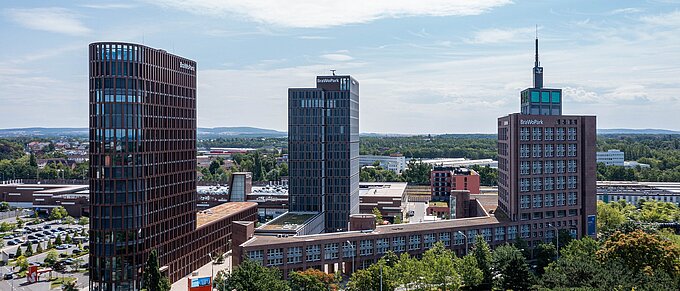Stadtansicht mit modernen Hochhäusern, grünen Bäumen und einem klaren Himmel im Hintergrund.