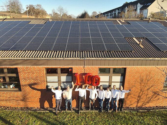 Gruppe von Menschen steht unter Solarzellen und feiert 100, mit roten Luftballons vor einem Backsteingebäude.