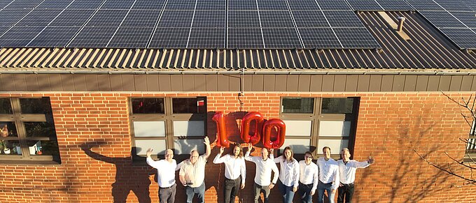 Gruppe von Menschen steht unter Solarzellen und feiert 100, mit roten Luftballons vor einem Backsteingebäude.