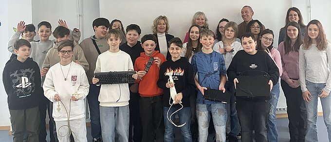 Eine Gruppe von Schülern und Lehrern posiert gemeinsam vor einer weißen Leinwand. Die Schüler halten zum Teil elektronische Gegenstände, wie Tastaturen, Fernbedienungen oder Mehrfachsteckdosen in den Händen.
