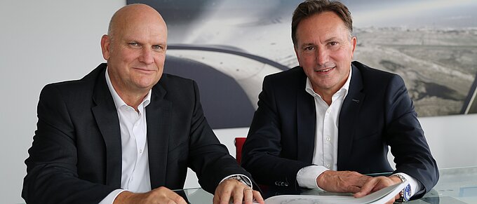 Ein Alternativtext für das Bild könnte sein: Zwei Männer in Anzügen sitzen an einem Glastisch, vor sich ein aufgeschlagenes Buch. Im Hintergrund hängt ein großes Bild, das eine Luftaufnahme einer Landschaft zeigt. Beide lächeln höflich in die Kamera.