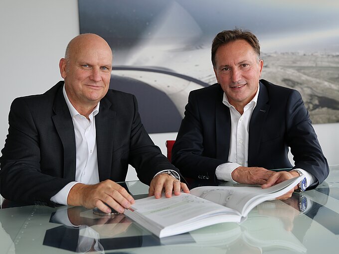 Ein Alternativtext für das Bild könnte sein: Zwei Männer in Anzügen sitzen an einem Glastisch, vor sich ein aufgeschlagenes Buch. Im Hintergrund hängt ein großes Bild, das eine Luftaufnahme einer Landschaft zeigt. Beide lächeln höflich in die Kamera.