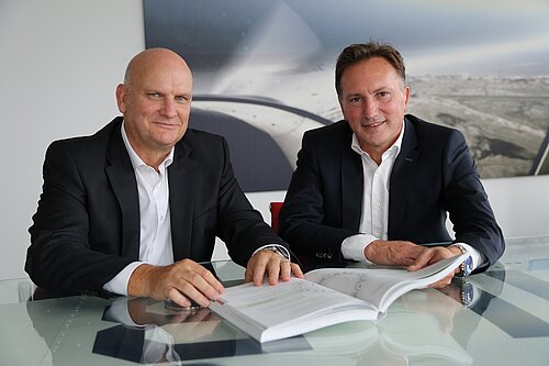 Ein Alternativtext für das Bild könnte sein: Zwei Männer in Anzügen sitzen an einem Glastisch, vor sich ein aufgeschlagenes Buch. Im Hintergrund hängt ein großes Bild, das eine Luftaufnahme einer Landschaft zeigt. Beide lächeln höflich in die Kamera.