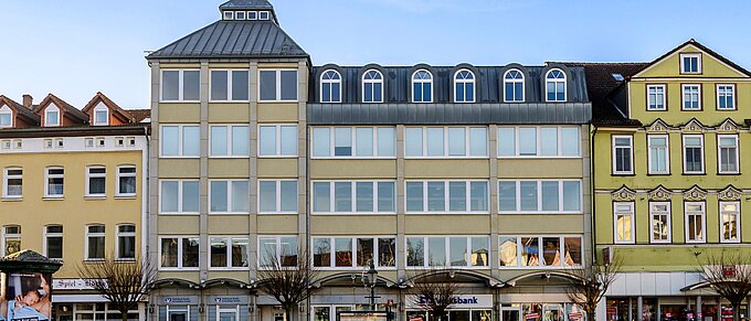 Städtische Häuserfassade mit Geschäften, kahlen Bäumen und einer Informationstafel auf einem sonnenbeschienenen Platz.