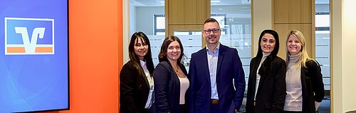 Fünf Personen in Bürokleidung lächeln in die Kamera, stehend vor einem Bildschirm mit Logo und oranger Wand.
