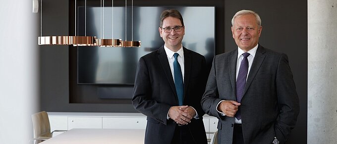 Zwei Männer in Anzügen stehen in einem modernen Büro mit einer großen Bildschirmpräsentation im Hintergrund.