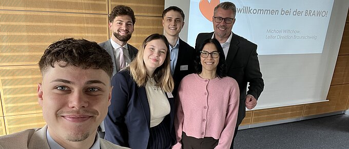 Gruppe von sechs Personen posiert lächelnd vor einer Präsentationstafel mit der Aufschrift "Willkommen bei der BRAWO!"
