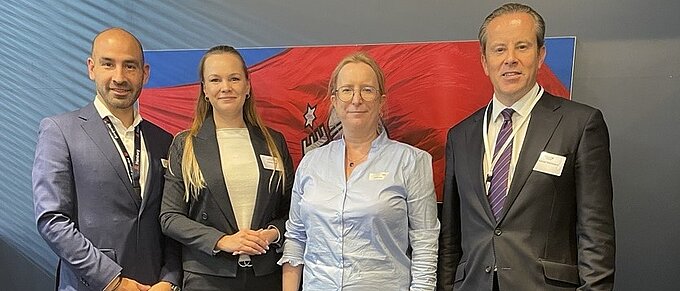 Vier Personen in Geschäftskleidung posieren vor einem roten Banner.