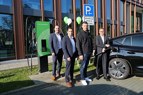 Vier Männer bei der Eröffnung einer neuen E-Ladestation neben einem schwarzen Auto und grüner "Bravo Green Energy"-Säule.