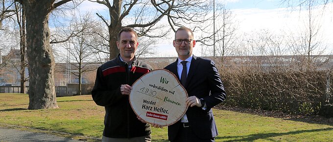 Zwei Männer halten ein großes rundes Holzschild mit der Aufschrift "Wir unterstützen mit 1.830 Bäumen. Werdet Harz Helfer. Jeder Baum zählt!"