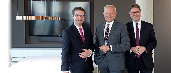 Drei Männer in Anzügen stehen lächelnd in einem modernen Konferenzraum mit Tisch und Bildschirm im Hintergrund.