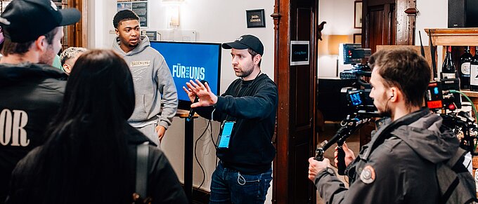 Filmcrew bei der Arbeit in einem Raum mit Jagdtrophäe an der Wand und einem Bildschirm, der "Für Uns" zeigt.