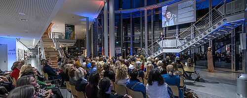 Ein Vortragender spricht zu einem großen Publikum in einem modernen, offenen Gebäude mit Treppenaufgang und Glaswänden.