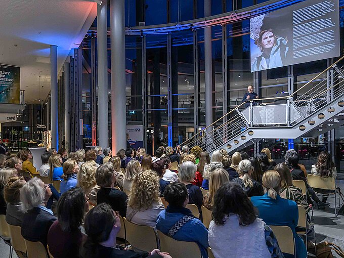 Ein Vortragender spricht zu einem großen Publikum in einem modernen, offenen Gebäude mit Treppenaufgang und Glaswänden.