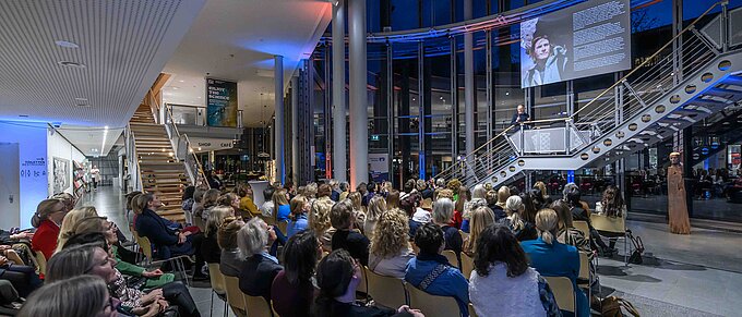 Ein Vortragender spricht zu einem großen Publikum in einem modernen, offenen Gebäude mit Treppenaufgang und Glaswänden.