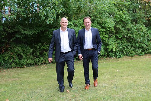 Zwei Männer in Anzügen gehen nebeneinander über eine Wiese. Sie tragen dunkle Anzüge mit weißen Hemden und wirken entspannt. Im Hintergrund ist dichtes, grünes Blattwerk zu sehen.