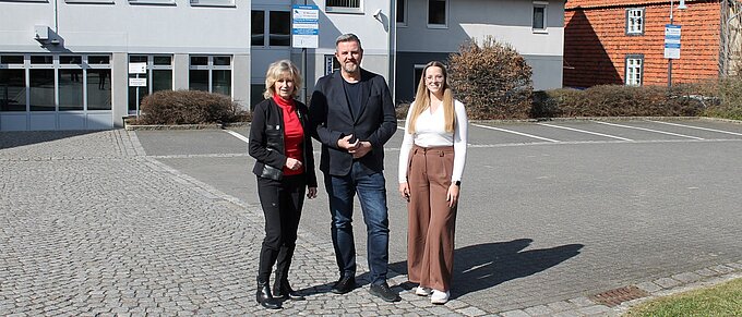 Drei Personen stehen lächelnd vor einem modernen Gebäude mit orangefarbenen Dächern an einem sonnigen Tag.