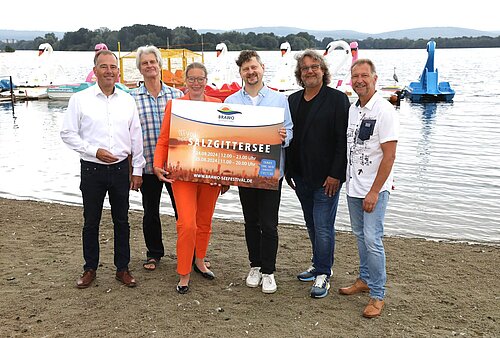 Die Verantwortlichen des BRAWO Seefests präsentieren am Strand des Salzgittersees das Plakat zur Ankündigung des Events.