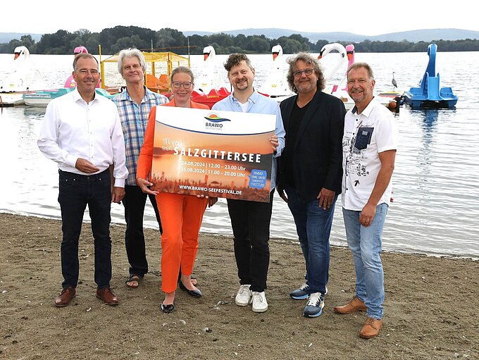 Die Verantwortlichen des BRAWO Seefests präsentieren am Strand des Salzgittersees das Plakat zur Ankündigung des Events.