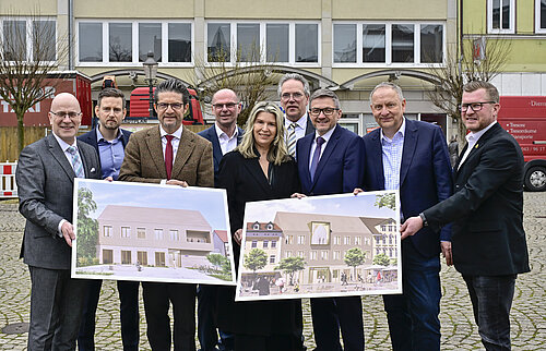 Gruppe von Personen präsentiert Architekturzeichnungen eines neuen Gebäudekomplexes auf einem öffentlichen Platz.