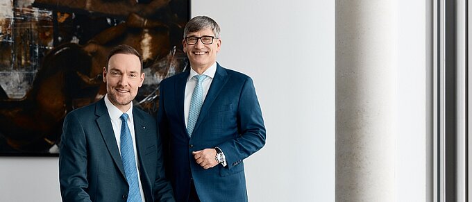 Zwei Männer in Anzügen posieren lächelnd in einem modernen Büro vor einem abstrakten Gemälde.
